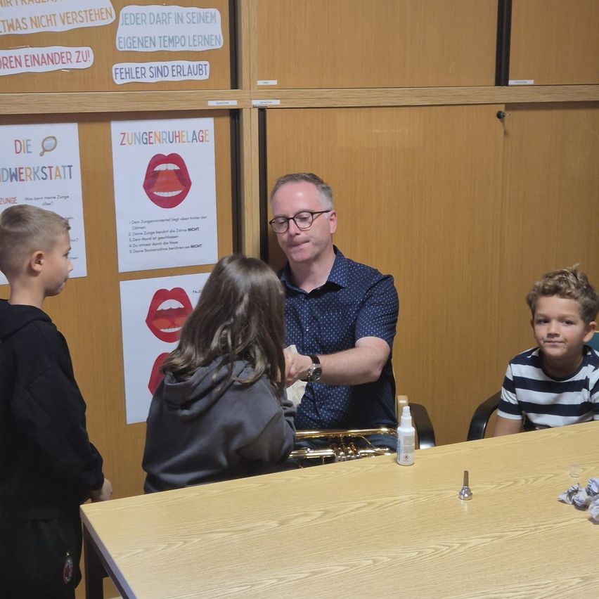 Ein Mann sitzt an einem Tisch mit drei Kindern. Er lächelt und hält ein Posaune. Die Kinder schauen ihn an und scheinen zuzuhören. Hinter ihnen ist eine Wand mit Plakaten.