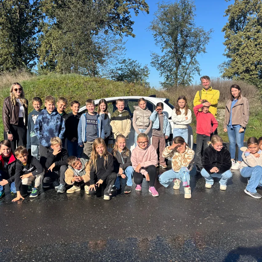 Eine Gruppe von Kindern und Erwachsenen posiert für ein Foto auf einer Straße mit einem weißen Auto im Hintergrund.