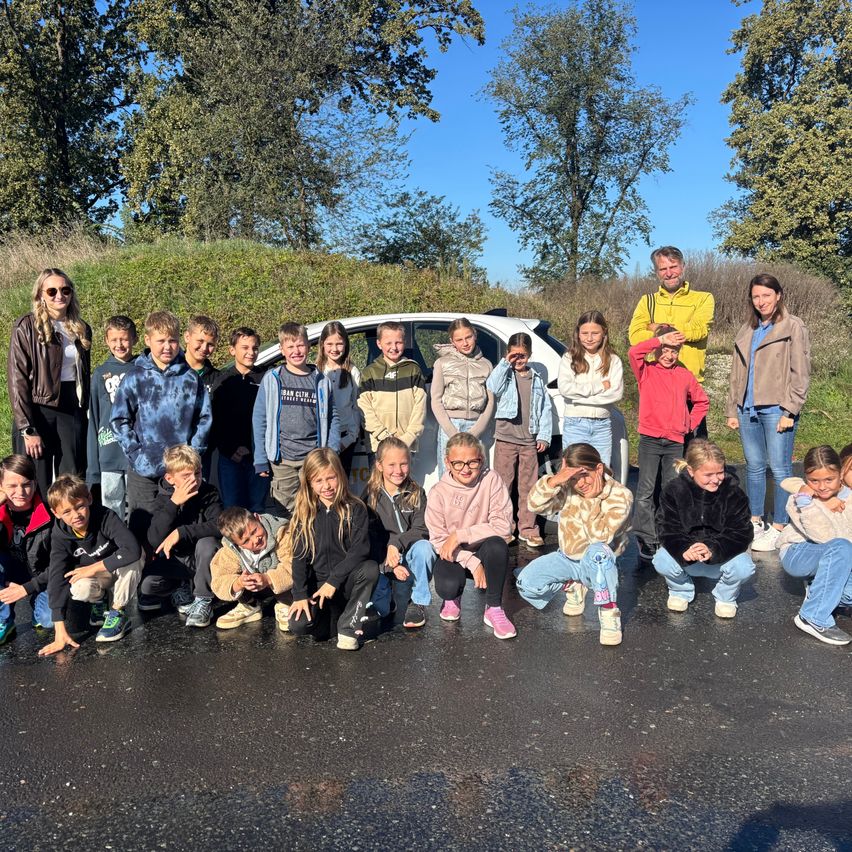 Eine Gruppe von Kindern und Erwachsenen posiert für ein Foto auf einer Straße mit einem weißen Auto im Hintergrund.