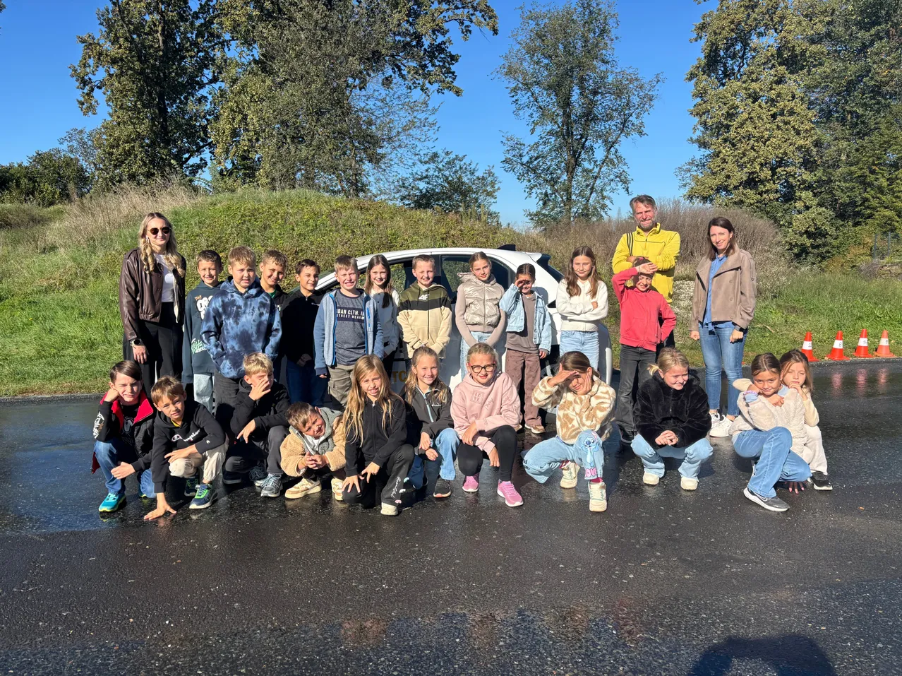 Eine Gruppe von Kindern und Erwachsenen posiert für ein Foto auf einer Straße mit einem weißen Auto im Hintergrund.