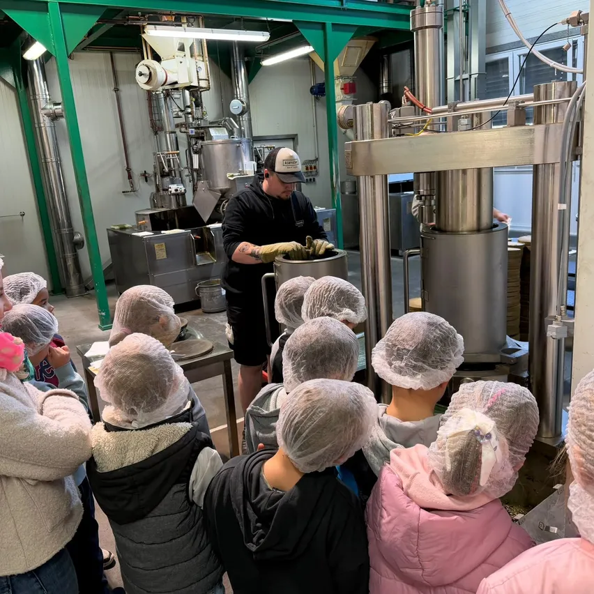 Eine Gruppe von Kindern mit Haarnetzen beobachtet einen Mann in einer Fabrik. Der Mann trägt einen Hut und Handschuhe und arbeitet mit einer Maschine.