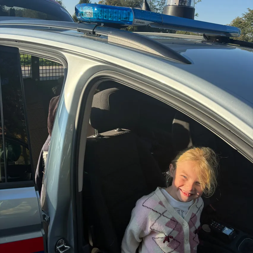 Ein kleines Mädchen mit blonden Haaren sitzt lächelnd in einem Polizeiauto.