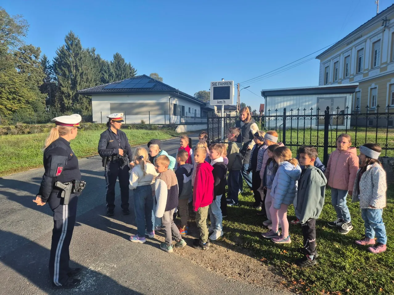 Eine Gruppe von Kindern steht auf dem Rasen vor einer Straße, wobei zwei Polizeibeamte am Straßenrand stehen.