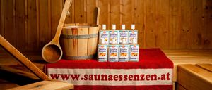 Ein roter Stoff mit dem Text 'www.saunaessenzen.at' bedeckt einen Holztisch mit einem Eimer, einem hölzernen Löffel und sechs Flaschen Sauna Essenz.