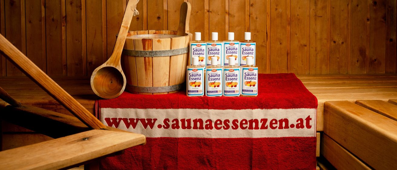 Ein Holzeimer, ein hölzerne Schöpfkelle und mehrere Flaschen Sauna Essenz stehen auf einem Tisch mit einem roten Tuch in einem Saunaraum.