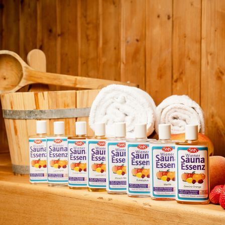 Mehrere Flaschen Sauna-Essenzen mit verschiedenen Aromen sind auf einer Holzoberfläche aufgereiht. Handtücher und Holzutensilien sind im Hintergrund.