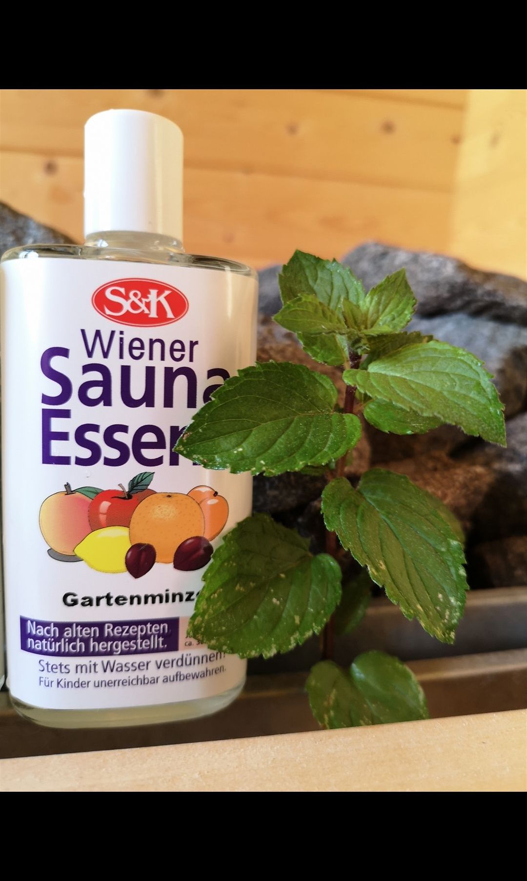Eine Flasche S&K Wiener Sauna Essigger Gartenminz steht neben einer Minzpflanze, umgeben von Steinen. Die Flasche ist mit Früchten beschriftet und hat ein rotes Logo.