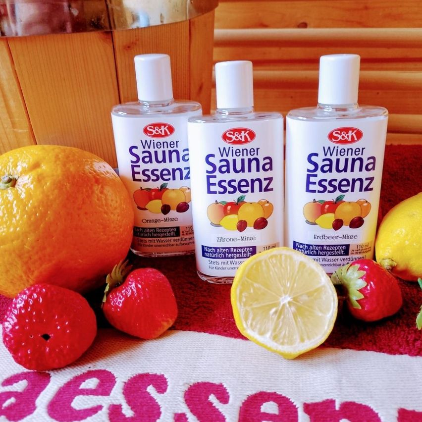 Drei Flaschen Wiener Sauna Essenz, eine Mischung aus Zitrus und Minze, sind zusammen mit einer geschnittenen Zitrone, einer ganzen Orange und mehreren Erdbeeren auf einem roten Tuch mit dem Wort 'Sauna' in Pink abgebildet.