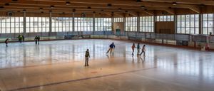 Eine überdachte Eisbahn mit Holzbalken und großen Fenstern. Mehrere Personen skaten auf dem Eis, einige in Gruppen und einige allein. Die Bahn ist gut beleuchtet mit mehreren an der Decke befestigten Leuchten.