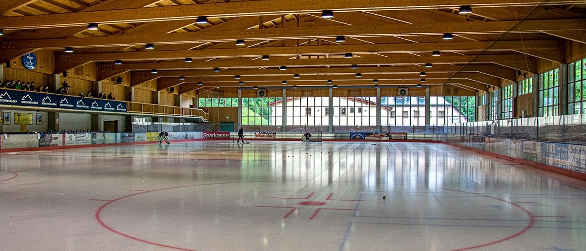 Eine überdachte Eishalle für Eishockey mit Holzdecke, Glaswänden und roten Markierungen auf dem Eis. Zwei Personen spielen Eishockey.