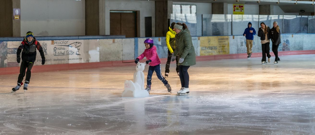 Indoor-Eisbahn mit Holzwänden und einem Banner. Eine Frau und ein Kind skaten in der Nähe eines Schneemanns, während zwei weitere Skater in der Nähe gehen.