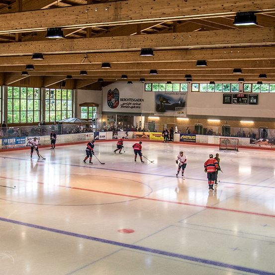 Eine überdachte Eishockeyhalle mit aktiven Spielern. Die Halle hat Holzdecken, Glaswände und Banner. Zuschauer sehen von den Tribünen zu.
