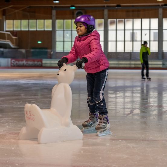 Ein junges Mädchen in einer rosa Jacke übt das Eislaufen in einer Eishalle und hält sich an einem weißen bärenförmigen Objekt fest. Sie trägt einen violetten Helm.