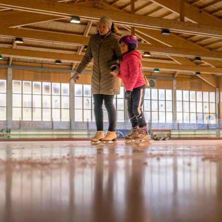 Eine Frau und ein Kind eislaufen in einer Halle. Die Frau trägt einen beigen Mantel und das Kind einen rosa Mantel und einen Helm. Sie eislaufen in der Nähe einer Glaswand.