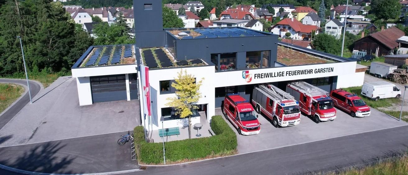 Luftaufnahme einer Feuerwache mit drei roten Feuerwehrautos vor dem Gebäude, einem Fahrrad, einer Fahne und Solarpanelen auf dem Dach.