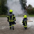 Zwei Feuerwehrleute in gelben und schwarzen Uniformen stehen vor einem Feuer. Einer hält einen Feuerlöscher und der andere beobachtet das Feuer.