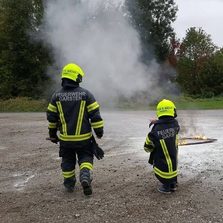 Zwei Feuerwehrleute in gelben und schwarzen Uniformen stehen vor einem Feuer. Einer hält einen Feuerlöscher und der andere beobachtet das Feuer.