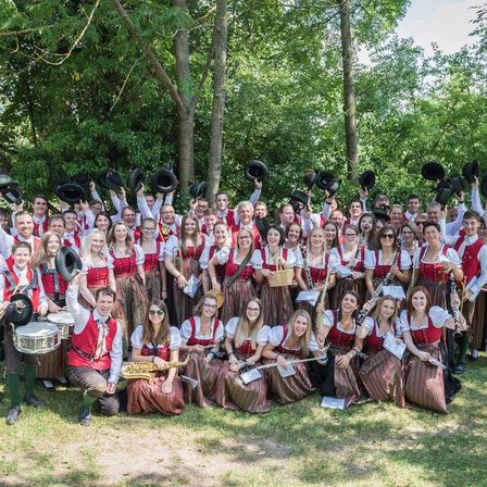 Eine große Gruppe von Musikern in traditioneller bayerischer Kleidung, einschließlich roter Westen und Dirndln, posiert für ein Foto in einem Park mit grünem Hintergrund. Sie halten Musikinstrumente wie Flöten, Klarinetten und Trommeln. Einige Mitglieder sitzen, während andere stehen und lächeln.