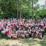 A large group of musicians in traditional Bavarian clothing, including red vests and dirndls, pose for a photo in a park with a green backdrop. They hold musical instruments such as flutes, clarinets, and drums. Some members are seated while others stand and smile.