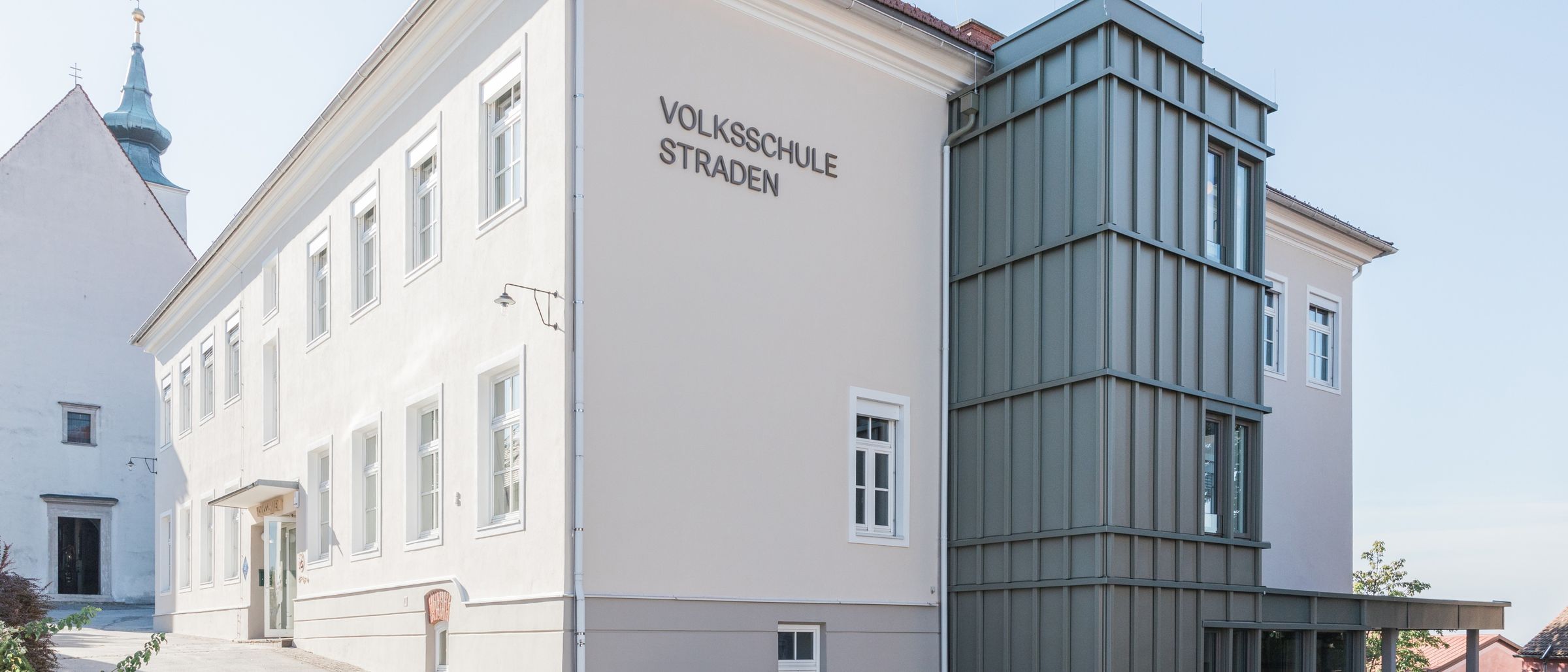 Ein weißes Gebäude mit mehreren Fenstern und einem Schild, das 'Volksschule Straden' auf der Seite steht. Es hat einen modernen grauen Anbau auf der rechten Seite.