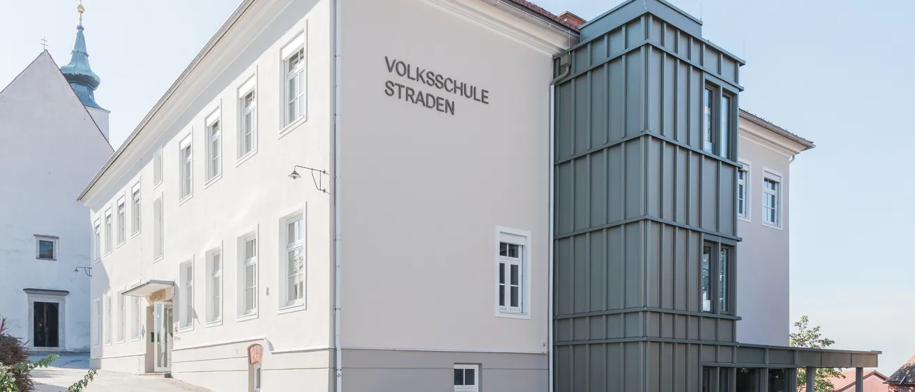 Ein weißes Gebäude mit mehreren Fenstern und einem Schild, das 'Volksschule Straden' auf der Seite steht. Es hat einen modernen grauen Anbau auf der rechten Seite.