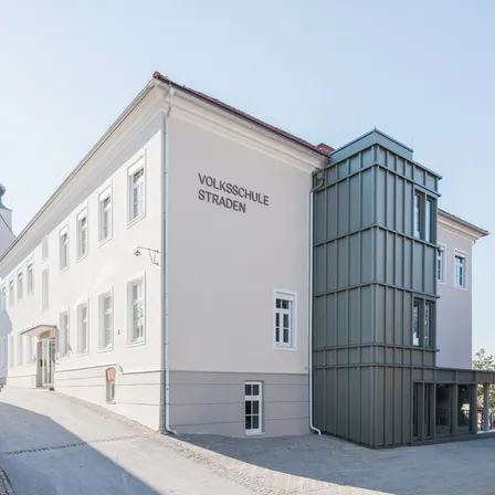 Ein weißes Gebäude mit mehreren Fenstern und einer grauen Erweiterung auf der rechten Seite. Das Gebäude trägt die Aufschrift 'Volksschule Straden'.