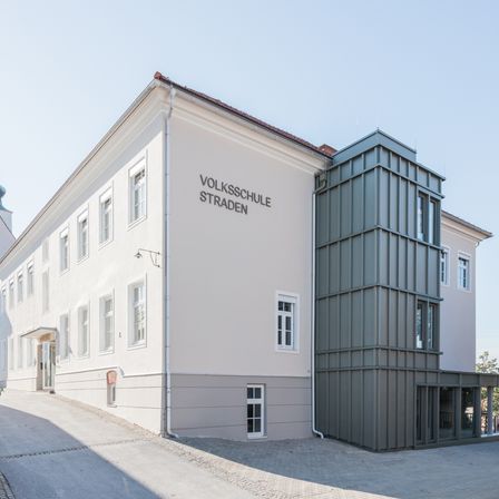 Ein weißes Gebäude mit mehreren Fenstern und einer grauen Erweiterung auf der rechten Seite. Das Gebäude trägt die Aufschrift 'Volksschule Straden'.