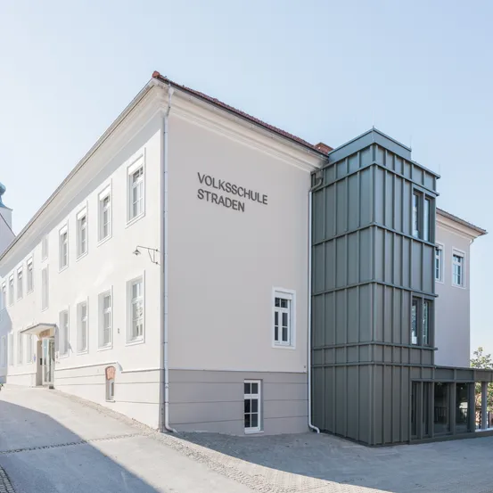 Ein weißes Gebäude mit mehreren Fenstern und einer grauen Erweiterung auf der rechten Seite. Das Gebäude trägt die Aufschrift 'Volksschule Straden'.