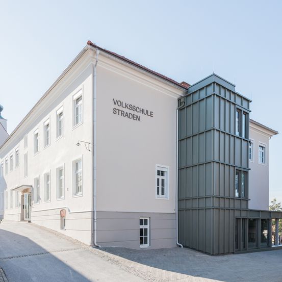 Ein weißes Gebäude mit mehreren Fenstern und einer grauen Erweiterung auf der rechten Seite. Das Gebäude trägt die Aufschrift 'Volksschule Straden'.