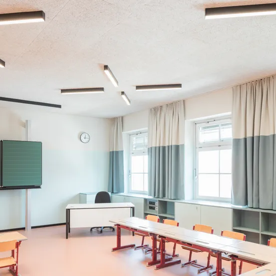 Ein leerer Klassenraum mit weißen Wänden, einer Tafel und Reihen von Tischen und Stühlen. Der Raum hat moderne Beleuchtung und Vorhänge.