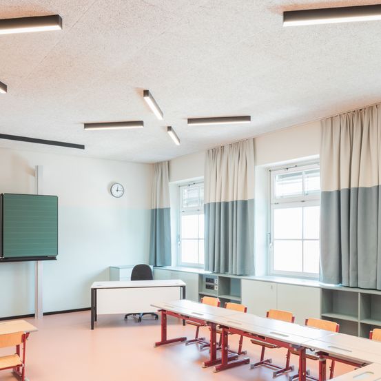 Ein leerer Klassenraum mit weißen Wänden, einer Tafel und Reihen von Tischen und Stühlen. Der Raum hat moderne Beleuchtung und Vorhänge.