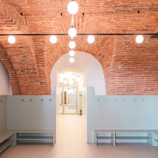 Ein leerer Raum mit Ziegelwänden, weißer Decke und einer Reihe von Haken an der Wand. Es gibt zwei Bänke auf beiden Seiten des Raumes.