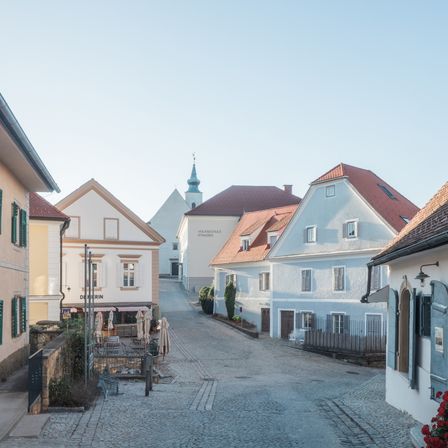 Eine Kopfsteinpflasterstraße gesäumt von traditionellen Gebäuden mit roten Dächern, eines davon hat ein Schild mit der Aufschrift 'Derrin'. Der Himmel ist klar und blau.