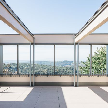 Ein moderner Balkon mit großen Fenstern und Blick auf eine hügelige Landschaft. Der Balkon hat einen gefliesten Boden und Metallgeländer.