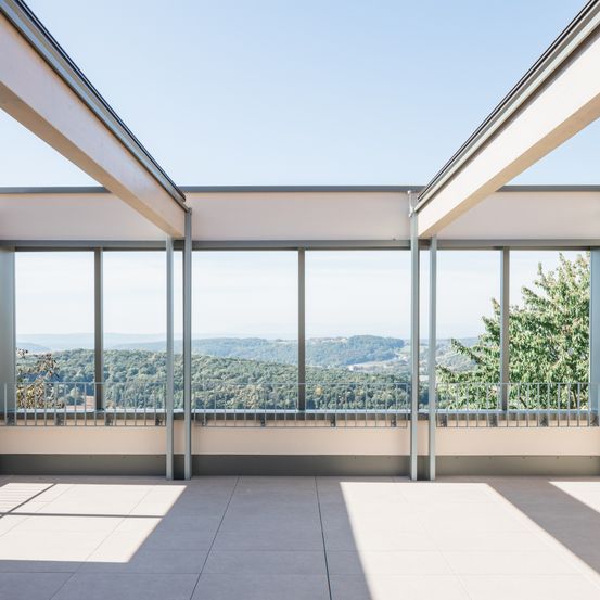 Ein moderner Balkon mit großen Fenstern und Blick auf eine hügelige Landschaft. Der Balkon hat einen gefliesten Boden und Metallgeländer.