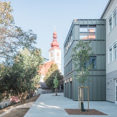 Ein modernes Gebäude mit grüner Fassade steht neben einer traditionellen Kirche mit rotem Dach und Turm. Das Gebiet ist von Bäumen umgeben und hat einen Betonweg.