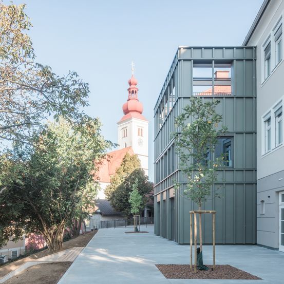Ein modernes Gebäude mit grüner Fassade steht neben einer traditionellen Kirche mit rotem Dach und Turm. Das Gebiet ist von Bäumen umgeben und hat einen Betonweg.