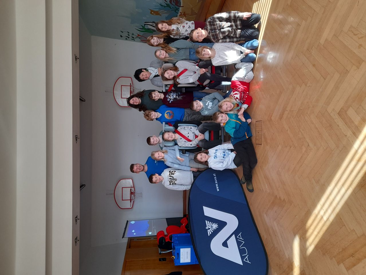 Eine Gruppe junger Menschen posiert für ein Foto, einige sitzen auf dem Boden, mit einem Basketballkorb im Hintergrund.