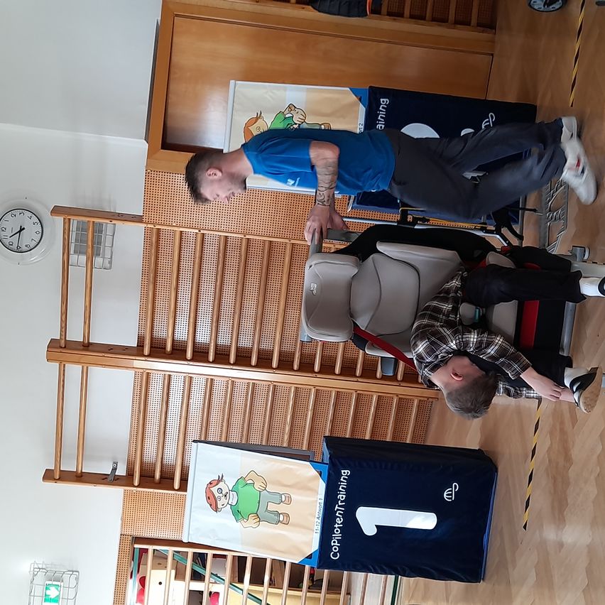 Ein Mann in einem blauen Shirt hilft einem Mann in einem karierten Shirt, der auf einem Stuhl sitzt. Sie befinden sich in einem Raum mit einer Uhr und Holzstufen.