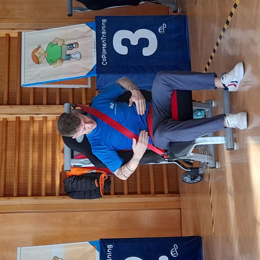 Ein Mann sitzt in einem Rollstuhl in einer Turnhalle, trägt ein blaues T-Shirt und einen roten Sicherheitsgurt. Im Hintergrund befinden sich blaue Banner mit der Nummer 3 und Text sowie eine hölzerne Treppe.