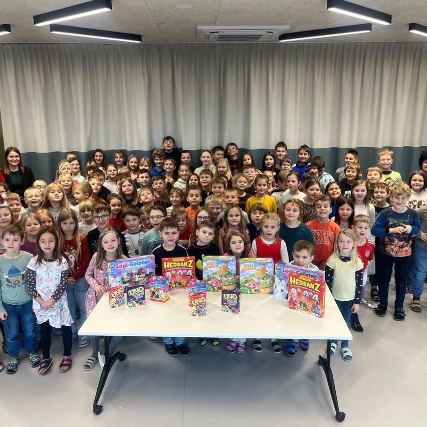 Ein Klassenzimmer voller Kinder, die lächelnd und für ein Foto posieren. Sie stehen um einen Tisch mit Brettspielen herum. Dahinter hängen weiße Vorhänge von der Decke.
