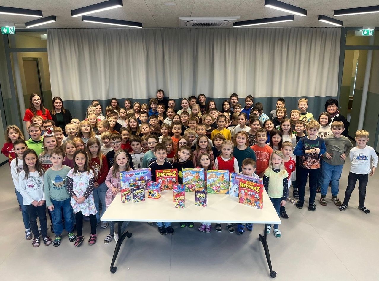 Ein Klassenzimmer voller Kinder, die lächelnd und für ein Foto posieren. Sie stehen um einen Tisch mit Brettspielen herum. Dahinter hängen weiße Vorhänge von der Decke.