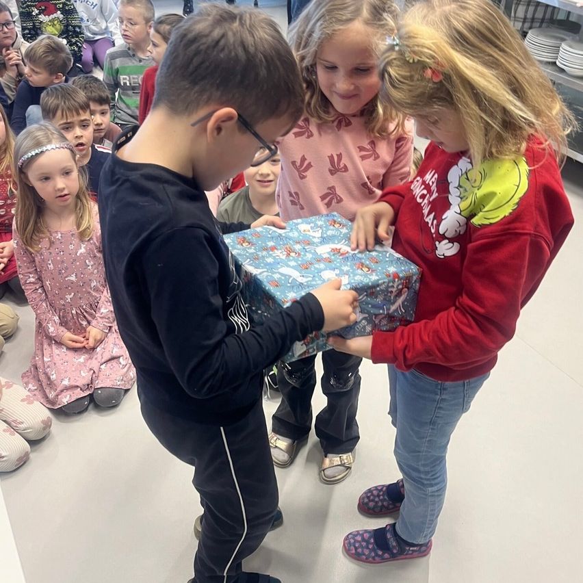 Ein Junge öffnet ein eingewickeltes Geschenk mit der Hilfe von zwei Mädchen, während andere Kinder sitzen und zusehen.