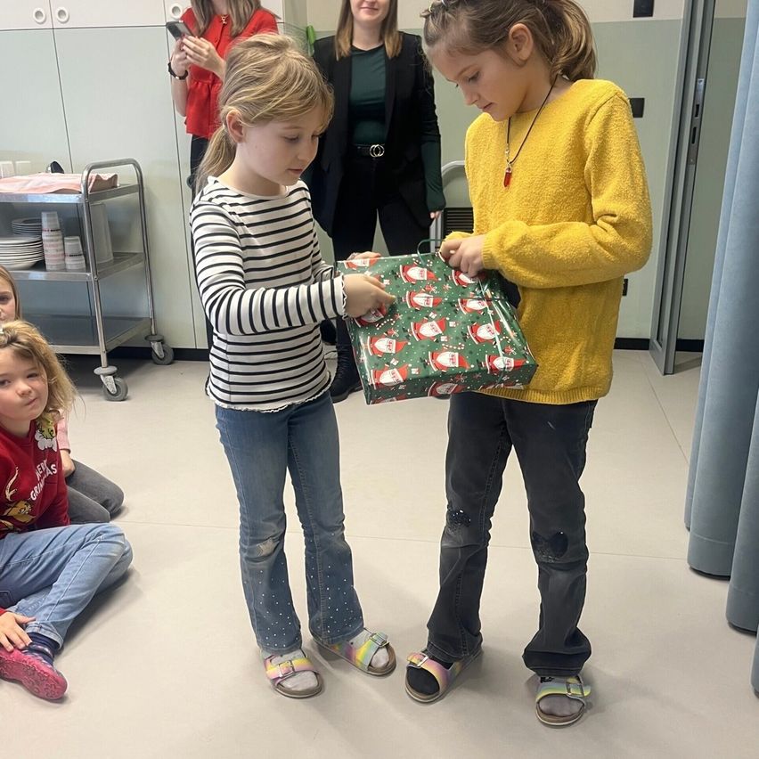 Zwei Mädchen in einem Krankenhaus-Setting tauschen Geschenke aus, eines der Mädchen hält ein eingewickeltes Geschenk. Hinter ihnen stehen eine Frau in Rot und eine in Grün in der Nähe eines Wagens. Ein Mädchen sitzt vor ihnen auf dem Boden.