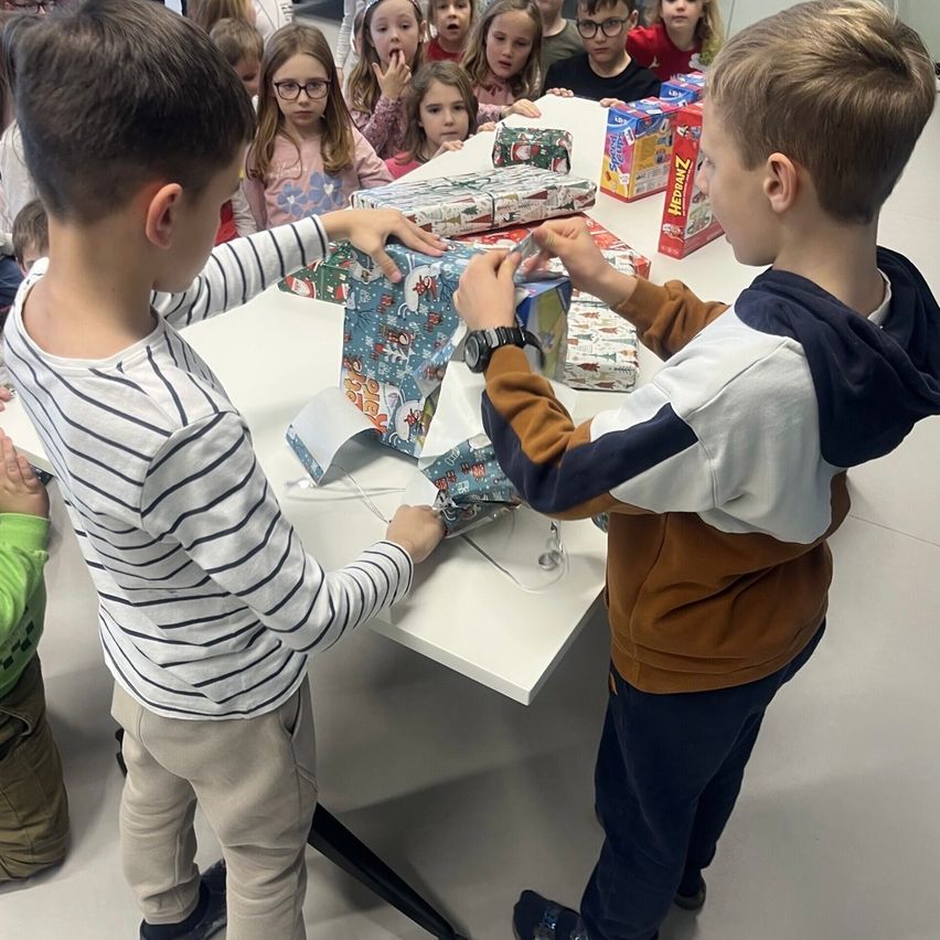 Zwei Jungen öffnen Geschenke auf einem Tisch, während andere Kinder zusehen. Ein Junge trägt ein gestreiftes Shirt und eine Hose, der andere ein Sweatshirt.