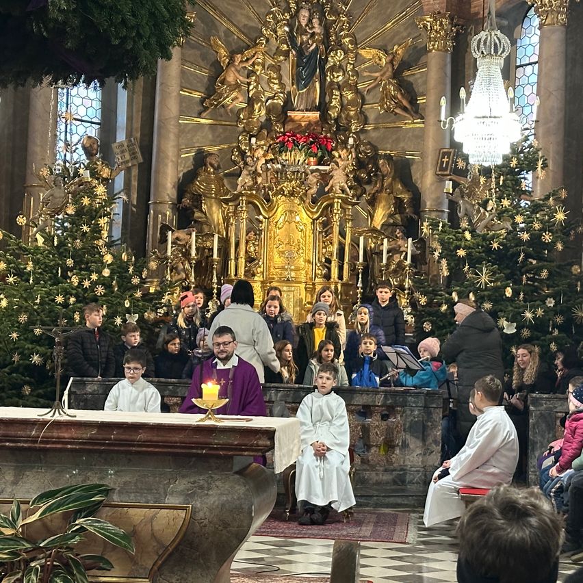 Ein Priester steht vor einem Altar mit einer brennenden Kerze, umgeben von Kindern in weißen Gewändern. Die Kirche ist mit Weihnachtsbäumen und Ornamenten geschmückt.