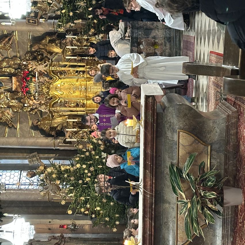 Eine Menschenmenge versammelt sich um einen Kirchenaltar, geschmückt mit goldenen Dekorationen und Weihnachtsbäumen. Priester und Teilnehmer sind anwesend, wobei ein Priester ein gelbes Band hält.