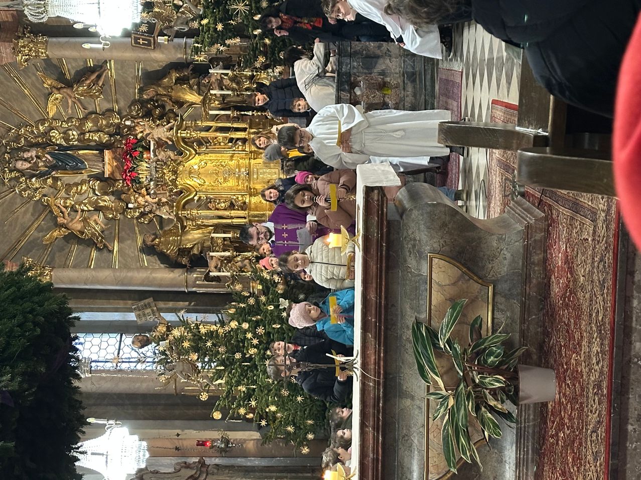 Eine Menschenmenge versammelt sich um einen Kirchenaltar, geschmückt mit goldenen Dekorationen und Weihnachtsbäumen. Priester und Teilnehmer sind anwesend, wobei ein Priester ein gelbes Band hält.
