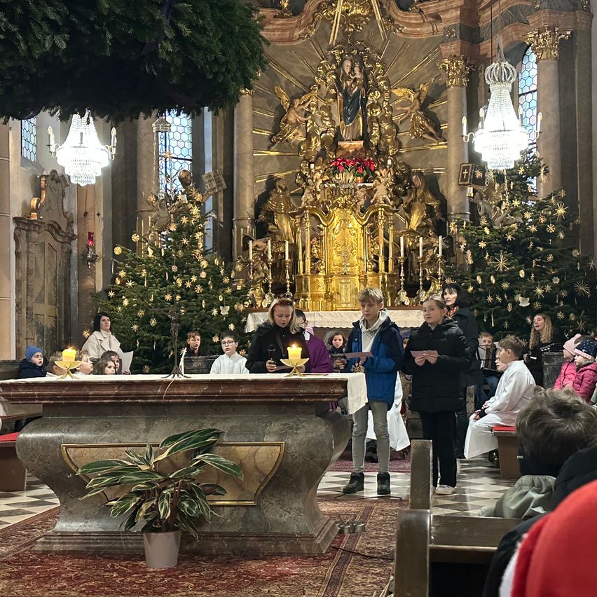 Eine Gruppe von Kindern steht während einer Zeremonie vor dem Altar in einer Kirche und hält Kerzen. Dahinter sind Weihnachtsbäume geschmückt.