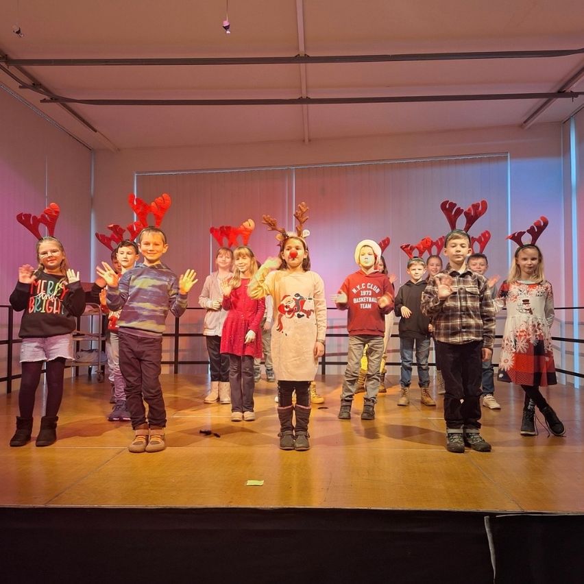 Eine Gruppe von Kindern mit roten Rentier-Kopfbändern und festlicher Kleidung steht auf der Bühne und singt oder spielt wahrscheinlich ein Weihnachtslied oder Theaterstück. Der Hintergrund ist eine Mischung aus rosa und blauen Farbtönen, wobei einige Kinder Turnschuhe und andere Stiefel tragen.